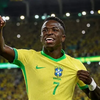Vini Jr. levanta o braço direito e puxa camisa da seleção, comemorando em partida, sorrindo. Imagem gerada por IA.