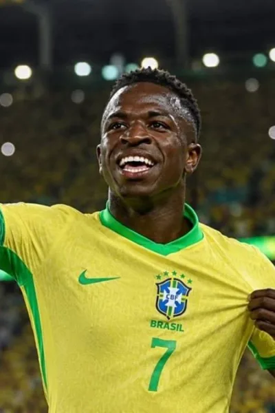 Vini Jr. levanta o braço direito e puxa camisa da seleção, comemorando em partida, sorrindo. Imagem gerada por IA.
