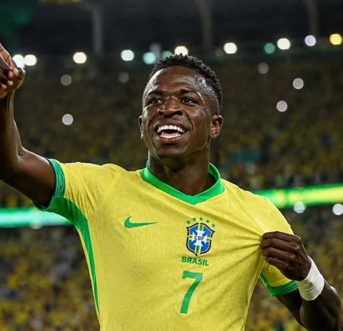 Vini Jr. levanta o braço direito e puxa camisa da seleção, comemorando em partida, sorrindo. Imagem gerada por IA.