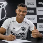 Vinicius Uchella, 20, assinando contrato com o Ceará Sporting Club, com a camisa do time e uma parede de coletiva de imprensa ao fundo. Imagem gerada por IA.