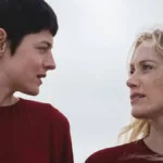 Esta imagem mostra Emma Corrin e Maika Monroe em uma cena do filme "100 Noites de Desejo" (título original: 100 Nights of Hero), protagonistas de vermelho se olham fixamente.