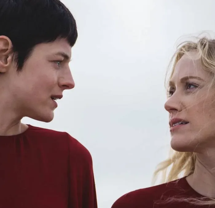 Esta imagem mostra Emma Corrin e Maika Monroe em uma cena do filme "100 Noites de Desejo" (título original: 100 Nights of Hero), protagonistas de vermelho se olham fixamente.