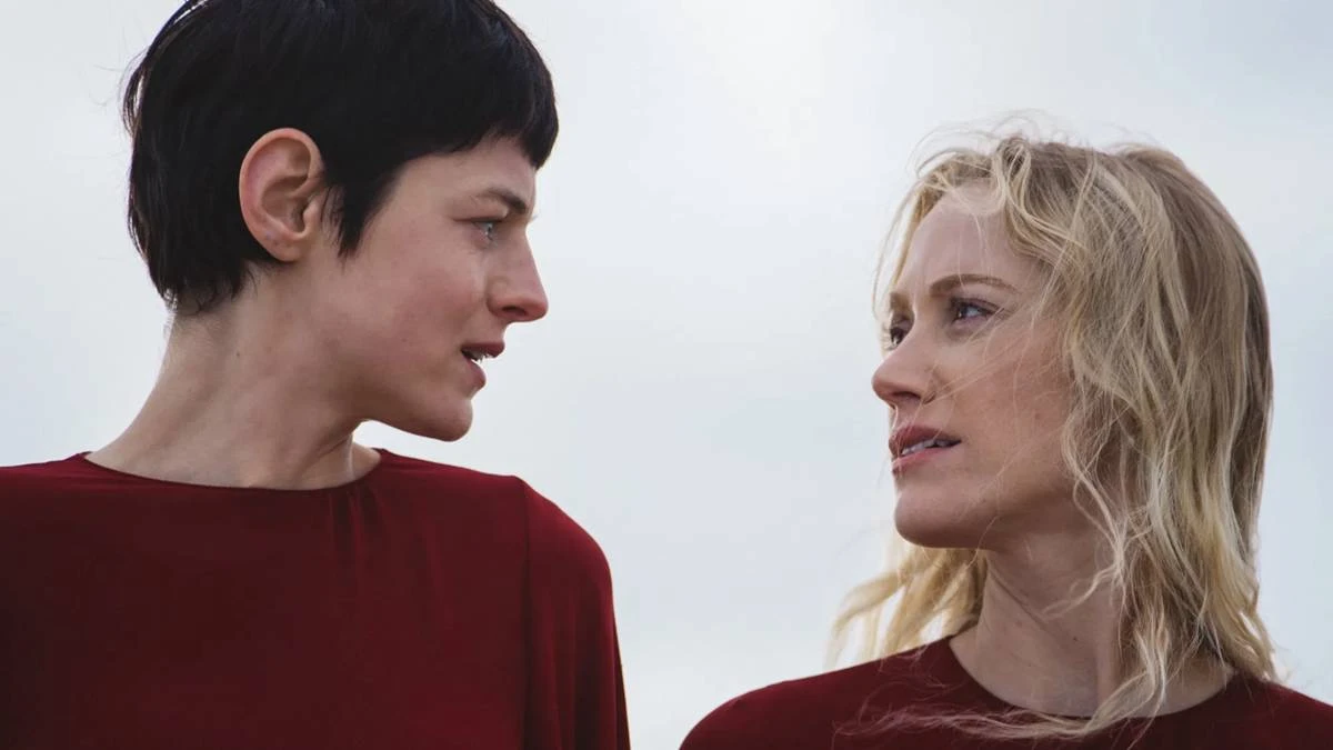 Esta imagem mostra Emma Corrin e Maika Monroe em uma cena do filme "100 Noites de Desejo" (título original: 100 Nights of Hero), protagonistas de vermelho se olham fixamente.