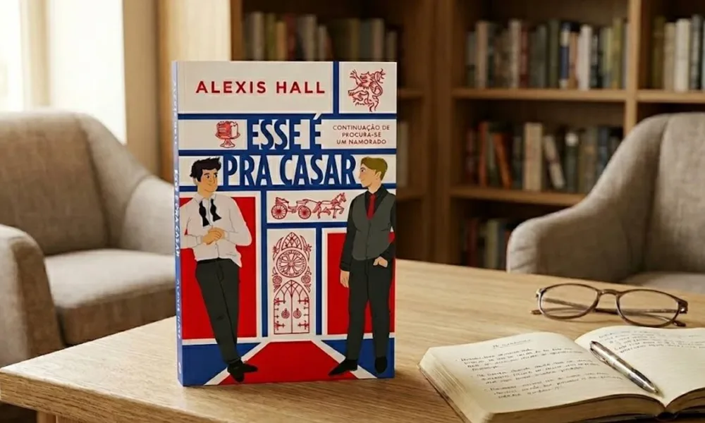 O livro na imagem é "Esse é pra casar", de Alexis Hall, de pé em um ambiente confortável de leitura, com uma estante ao fundo, luz ambiente diurna. Há um livro com anotações e caneta, e um óculos fechado na mesma mesa.