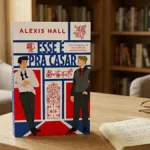 O livro na imagem é "Esse é pra casar", de Alexis Hall, de pé em um ambiente confortável de leitura, com uma estante ao fundo, luz ambiente diurna. Há um livro com anotações e caneta, e um óculos fechado na mesma mesa.