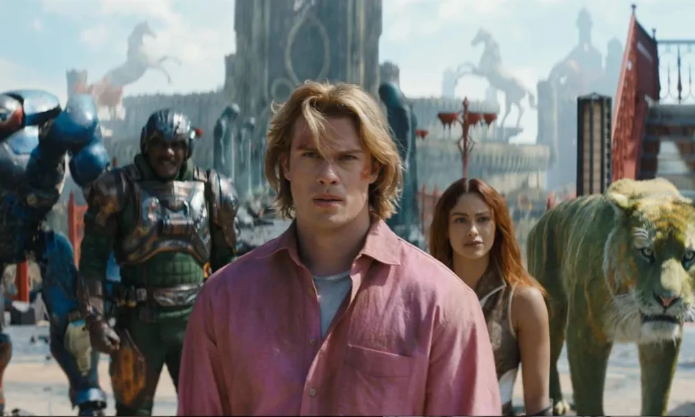 Esta é uma cena do filme live-action Mestres do Universo, com estreia prevista para junho de 2026. Adam está ao centro, camisa rosa, e o elenco de Eternia ao fundo.