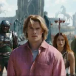 Esta é uma cena do filme live-action Mestres do Universo, com estreia prevista para junho de 2026. Adam está ao centro, camisa rosa, e o elenco de Eternia ao fundo.