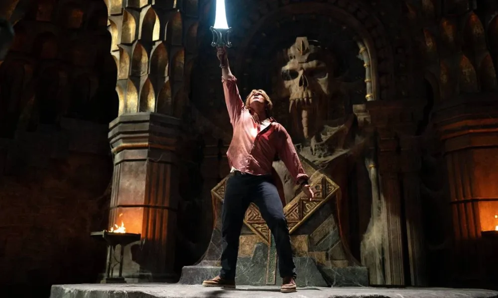 Esta é uma cena do filme live-action Mestres do Universo, estrelado por Nicholas Galitzine. A imagem mostra o protagonista em uma pose clássica, erguendo a Espada do Poder.