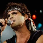 Cazuza em apresentação, câmera em suave contra plongée com artista segurando microfone no palco, fundo escuro iluminado. Imagem gerada por IA.