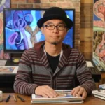 Shintaro Kago em estúdio próprio com suas obras ao lado, painel na parede mostra seu estilo artístico bem marcado no surreal. Ele está com chapéu-coco, óculos, expressão neutra. Imagem gerada por IA.