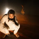 A Hora do Boi | Quando Existir Não Basta 13 Ator agachado no palco, iluminado por uma luz intensa vinda de trás, durante cena de A Hora do Boi.