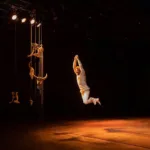 A Hora do Boi | Quando Existir Não Basta 14 Ator suspenso no ar durante movimento cênico no palco, próximo a cordas e elementos cenográficos, sob iluminação quente.