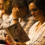 A Hora do Boi | Quando Existir Não Basta 15 Mulher de óculos lê o programa de A Hora do Boi enquanto outros espectadores aparecem sentados ao fundo.