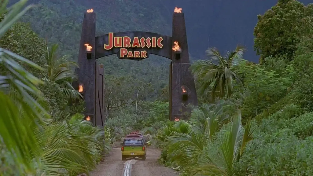 Entrada do Jurassic Park em meio a uma floresta tropical, com grandes portões de pedra e tochas acesas. Veículos percorrem a estrada que leva ao parque. Filme Jurassic park
